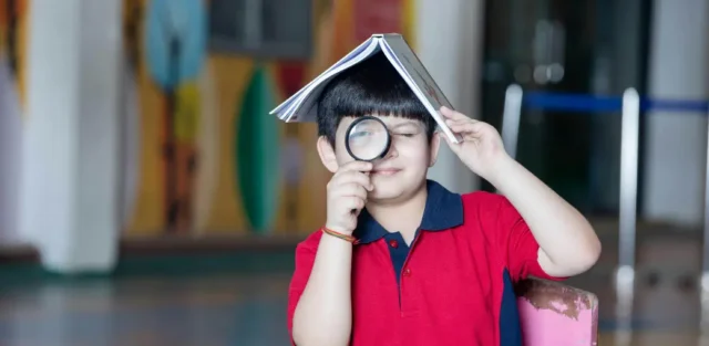 A young child playfully holding a magnifying glass to their eye with a book balanced on their head, illustrating the unique perspective and potential of a child with high-needs autism.