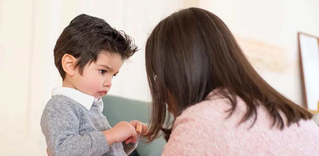 Young boy with dark hair looking down upset while his mother leans in close to comfort him